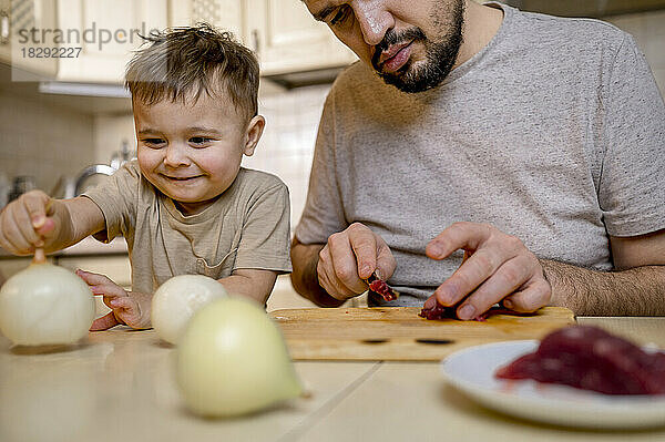 Lächelnder Junge nimmt weiße Zwiebeln  während sein Vater in der Küche rotes Fleisch schneidet