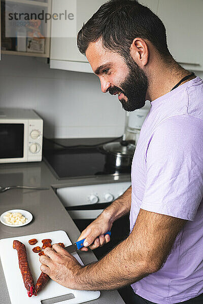 Lächelnder Mann schneidet Chorizo-Wurst an der Küchentheke