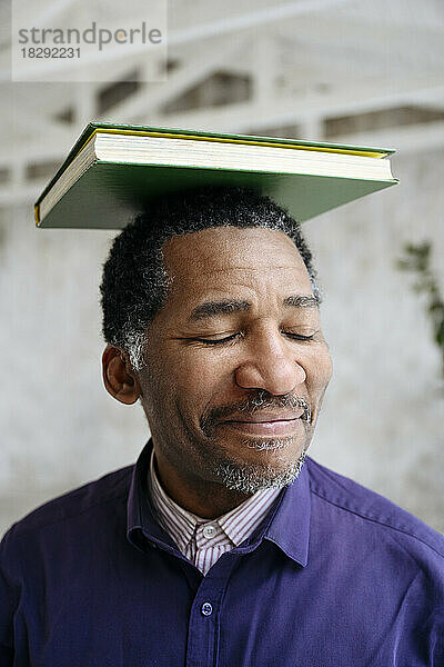 Lächelnder reifer Mann mit geschlossenen Augen  der ein Buch auf dem Kopf balanciert