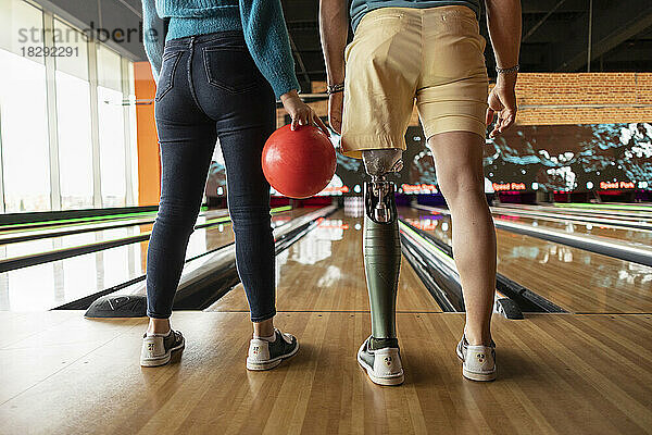 Frau hält Ball mit Freund  der künstliche Gliedmaßen trägt und auf Hartholzboden in der Bowlingbahn steht