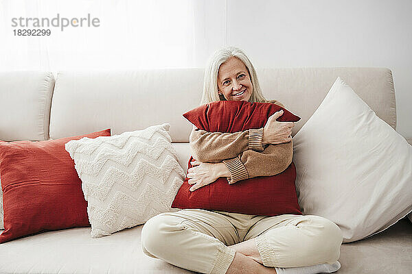 Glückliche reife Frau mit Kissen sitzt zu Hause auf dem Sofa
