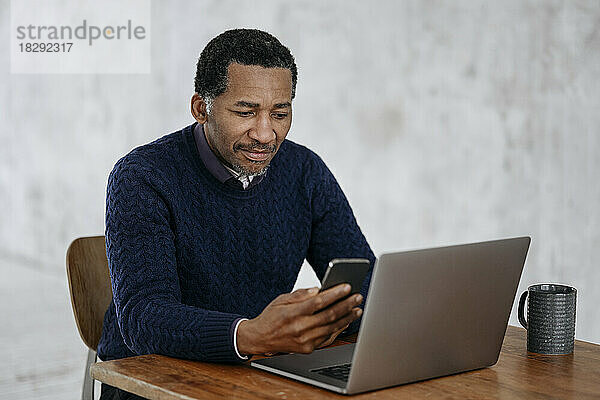 Geschäftsmann schaut auf sein Mobiltelefon und sitzt mit Laptop auf dem Tisch im Büro