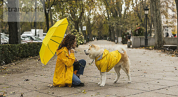 Junge Frau hält Regenschirm und kauert mit Hund am Fußweg