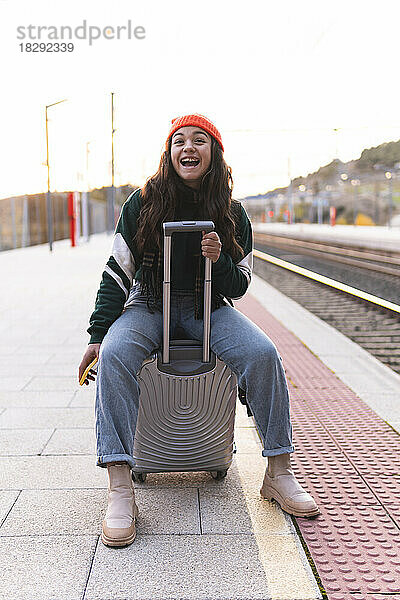 Fröhliches Mädchen mit Strickmütze sitzt auf Koffer am Bahnsteig