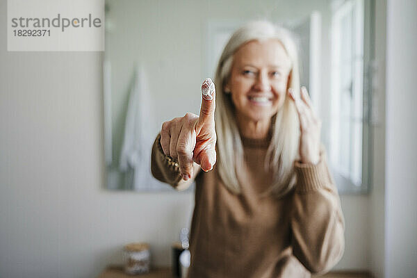 Lächelnde reife Frau mit Feuchtigkeitscreme am Finger im Badezimmer