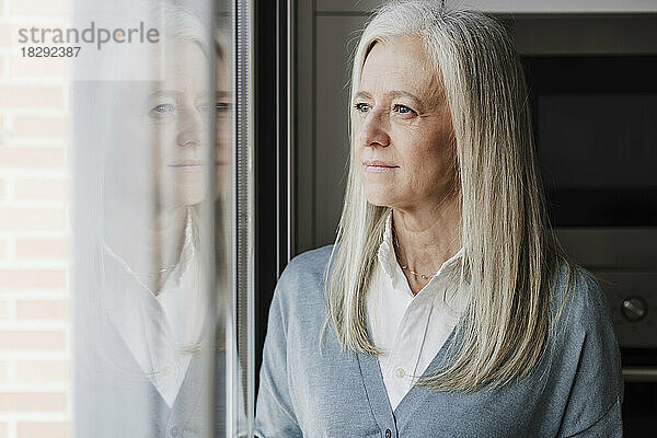 Nachdenkliche reife Frau mit grauen Haaren am Fenster zu Hause