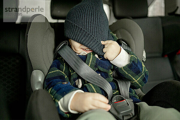 Fröhlicher Junge bedeckt Gesicht mit Strickmütze im Autositz
