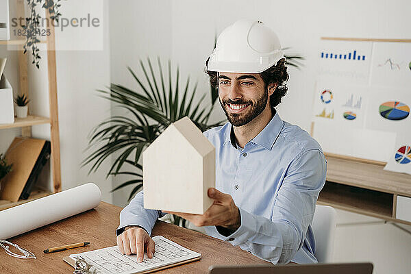 Lächelnder Architekt mit Bauplan hält Hausmodell am Schreibtisch im Büro