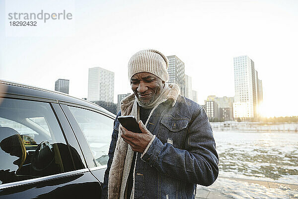 Lächelnder Mann benutzt im Winter sein Smartphone vor dem Auto