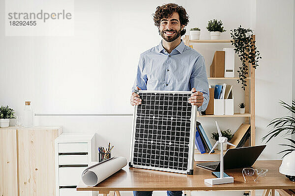 Glücklicher Geschäftsmann mit Solarpanel am Schreibtisch im Büro