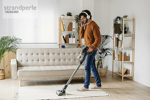 Glücklicher junger Mann mit kabellosen Kopfhörern  der sein Zuhause mit einem Staubsauger putzt