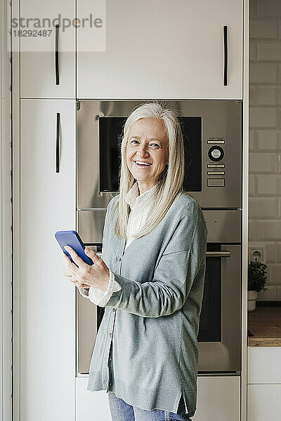 Glückliche reife Frau mit Smartphone  die in der Küche steht