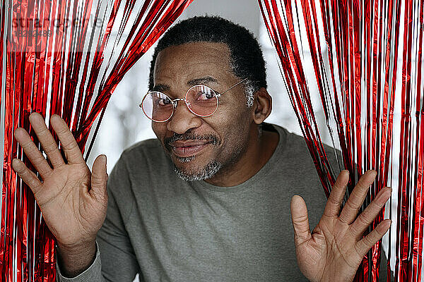 Happy mature man wearing pink glasses looking through red tinsel curtain