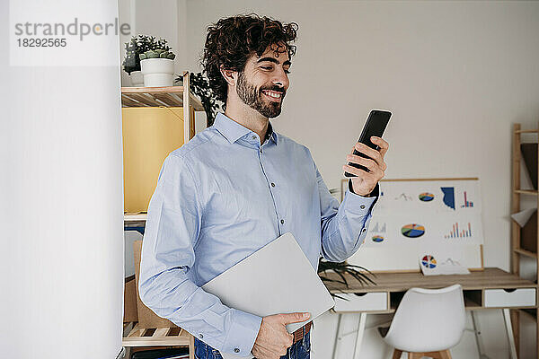Glücklicher junger Ingenieur  der im Büro sein Mobiltelefon nutzt und sein Smartphone in der Hand hält