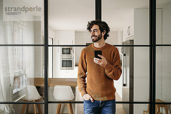 Glücklicher junger Mann mit Smartphone  der zu Hause an einer Glastür lehnt