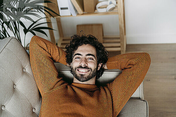 Glücklicher junger Mann entspannt sich zu Hause mit den Händen hinter dem Kopf auf dem Sofa