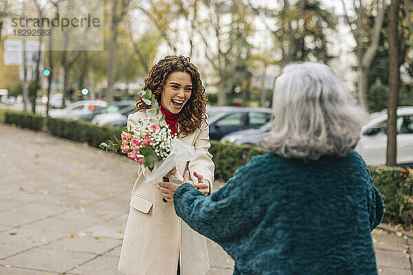 Fröhliche junge Frau schenkt Großmutter am Fußweg einen Blumenstrauß