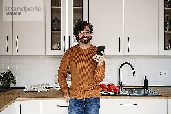 Glücklicher Mann mit Smartphone  der zu Hause auf der Küchentheke lehnt