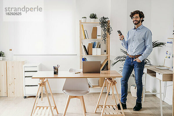 Lächelnder junger Geschäftsmann steht mit Smartphone im Büro