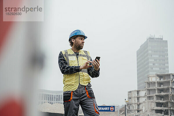Lächelnder reifer Arbeiter mit Smartphone auf der Baustelle