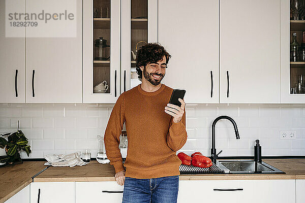 Happy man using smart phone leaning on kitchen counter at home