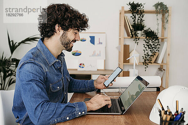 Lächelnder Ingenieur  der an Laptop und Smartphone arbeitet und am Schreibtisch im Büro sitzt
