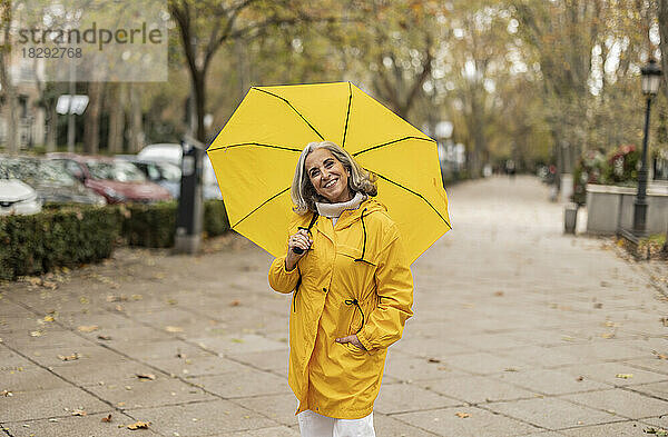 Glückliche ältere Frau mit gelbem Regenschirm steht auf dem Fußweg