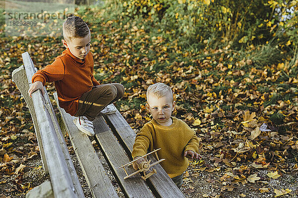 Junge schaut Bruder an  der im Herbst mit Spielzeugflugzeug auf Bank spielt