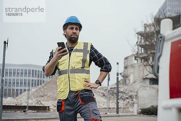 Arbeiter hält Smartphone mit der Hand auf der Hüfte vor der Baustelle