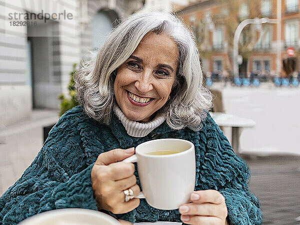 Glückliche ältere Frau mit Kaffeetasse sitzt im Straßencafé