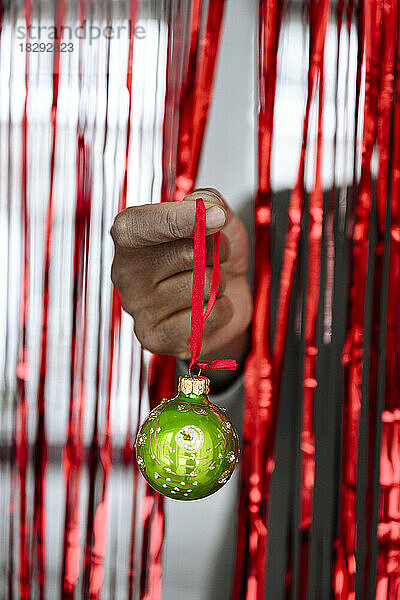 Hand of man holding green Christmas bauble in front of red tinsel curtain