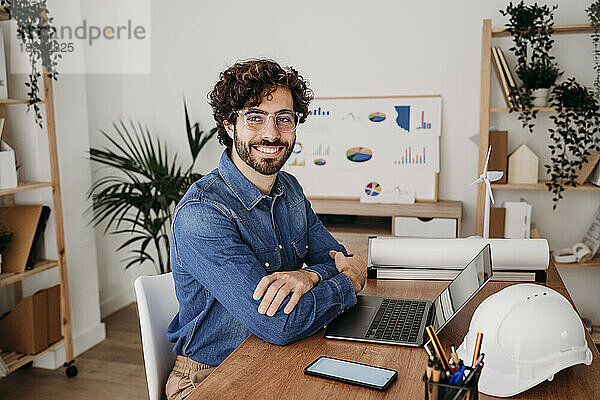 Glücklicher junger Ingenieur mit Brille sitzt mit Laptop am Schreibtisch im Büro