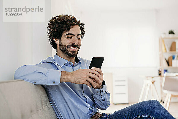 Glücklicher junger Geschäftsmann sitzt mit Smartphone auf dem Sofa im Büro