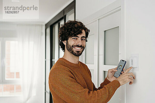 Glücklicher junger Mann  der zu Hause Thermostat mit Smartphone bedient