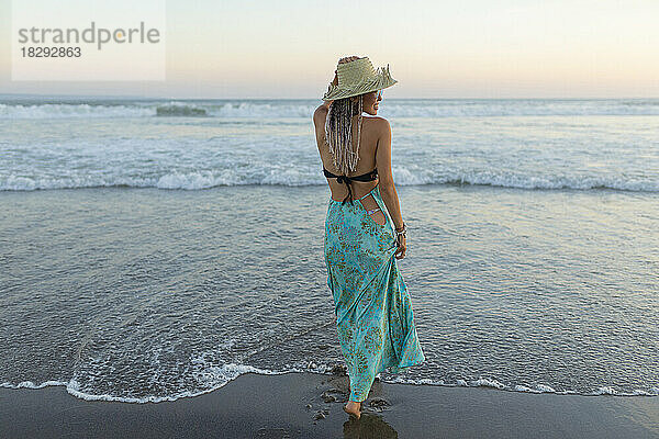 Junge Frau mit Hut steht am Strand vor dem Meer