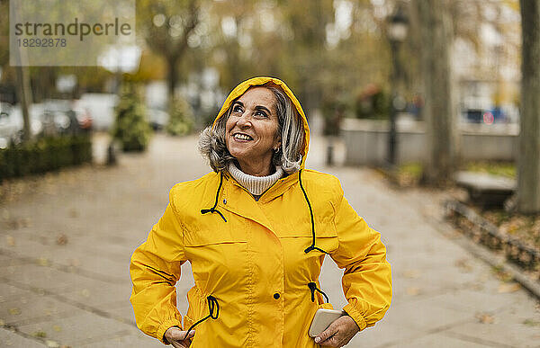 Nachdenkliche ältere Frau im gelben Regenmantel steht am Fußweg