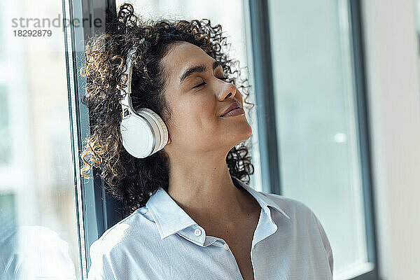 Lächelnde Frau mit kabellosen Kopfhörern lehnt zu Hause am Fenster