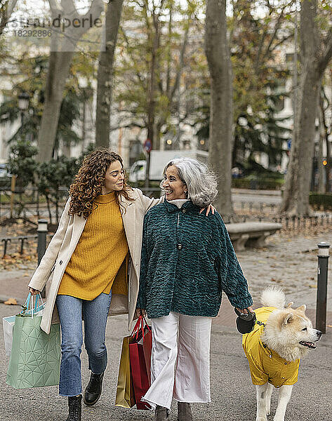 Glückliche Enkelin und Großmutter halten Einkaufstüten in der Hand und gehen mit Hund am Fußweg spazieren