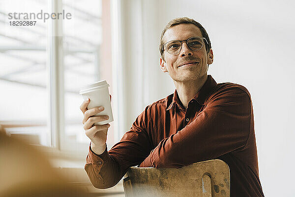 Lächelnder Geschäftsmann mit Einwegkaffeetasse sitzt im Bürostuhl