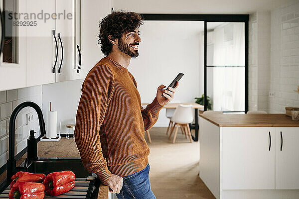 Glücklicher junger Mann steht mit Handy zu Hause an der Küchentheke