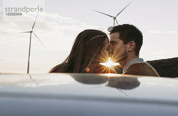 Couple kissing by car at sunset with wind farm in background