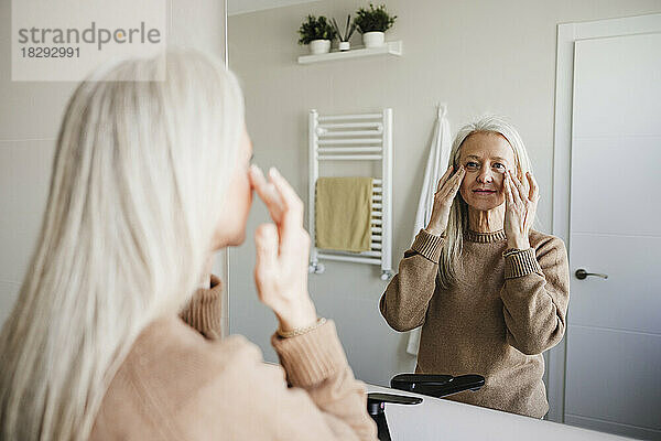 Reife Frau blickt in den Spiegel und trägt Feuchtigkeitscreme auf ihr Gesicht auf