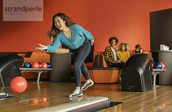 Young woman throwing ball with friends sitting in background at bowling alley