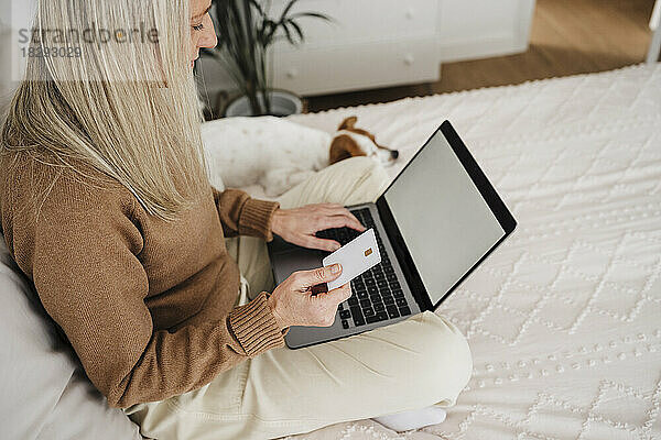 Reife Frau bezahlt mit Kreditkarte über Laptop im Schlafzimmer
