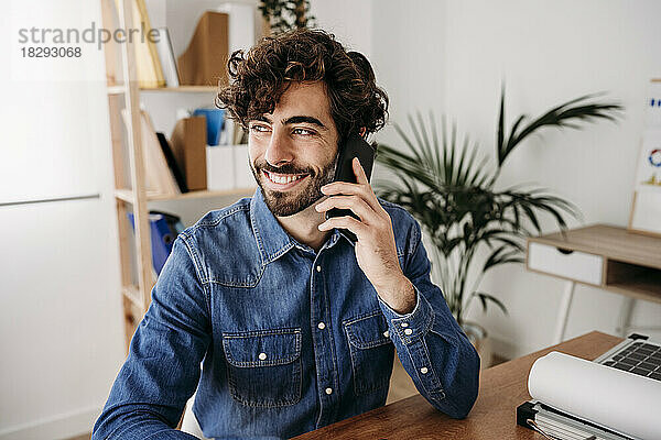 Glücklicher junger Ingenieur  der am Schreibtisch im Büro sitzt und mit dem Mobiltelefon spricht
