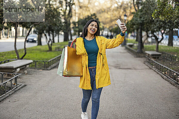 Glückliche Frau mit Einkaufstüten  die auf dem Fußweg ein Selfie mit dem Handy macht