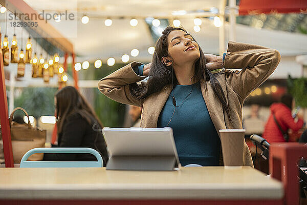 Lächelnde junge Frau mit den Händen hinter dem Kopf sitzt im Café