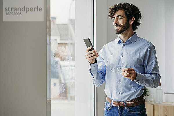 Glücklicher Geschäftsmann mit Smartphone  der durch das Fenster im Büro schaut