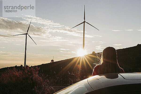 Frau lehnt an Auto und blickt bei Sonnenuntergang auf Windpark