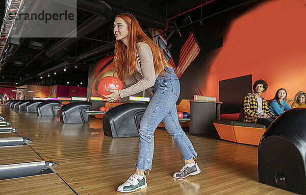 Junge Frau wirft Ball mit Freunden  die im Hintergrund auf der Bowlingbahn sitzen